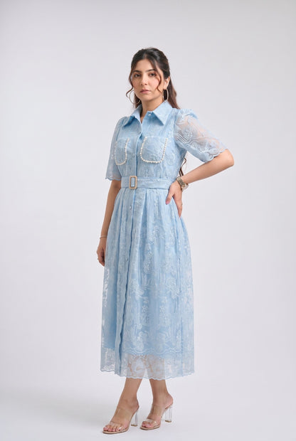 A woman modeling a light blue midi dress with sheer short sleeves, a pointed collar, and a matching belt with a decorative gold-tone buckle, photographed against a plain light gray studio background.