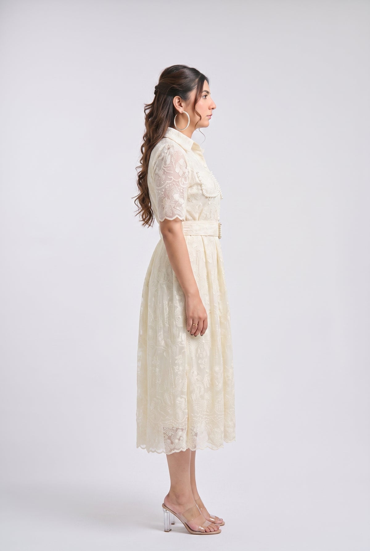 A woman stands in profile wearing a cream lace midi dress with short sleeves, a collar, and a matching belt, photographed against a plain light gray studio background.