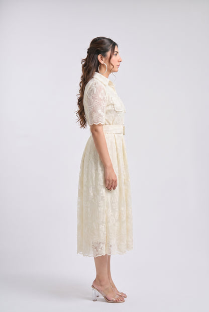 A woman stands in profile wearing a cream lace midi dress with short sleeves, a collar, and a matching belt, photographed against a plain light gray studio background.