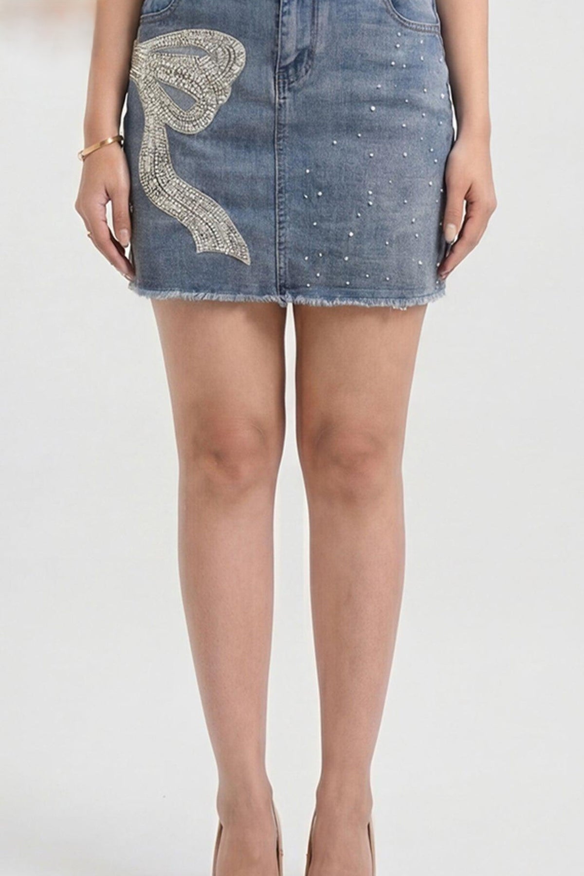 A model's lower body wearing a light-wash blue denim mini skirt with a large silver rhinestone bow applique on the left side and small silver rhinestone embellishments scattered across the front.