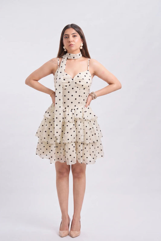 A woman modeling a sleeveless halter-neck mini dress with a tiered ruffle skirt in a cream color with black polka dots, paired with a matching choker and beige heels, against a light gray studio background.