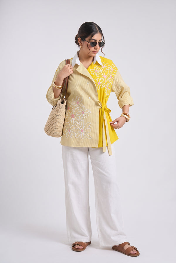 A woman modeling a light yellow embroidered shirt with a white collar, paired with white wide-leg pants, brown sandals, and a beige woven shoulder bag against a light gray studio background.