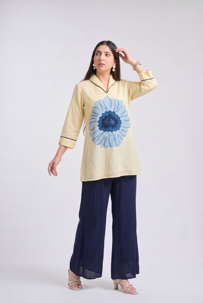A woman modeling a light yellow tunic top with a large blue flower print on the front, paired with dark navy wide-leg pants, against a light gray studio background.