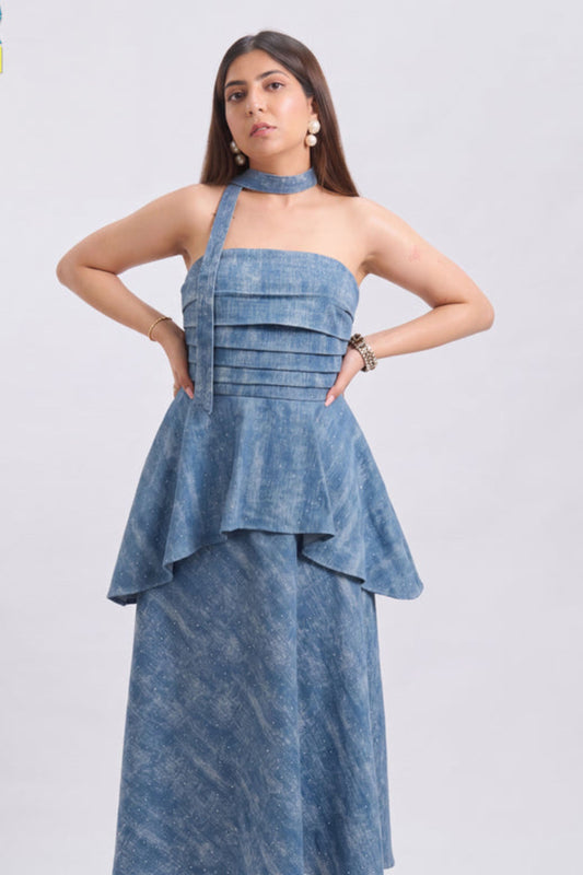 A woman modeling a blue halter-neck maxi dress with a textured, washed denim-like finish. The dress features a structured, ruched bodice with horizontal pleats, a peplum waist, and a matching long skirt.