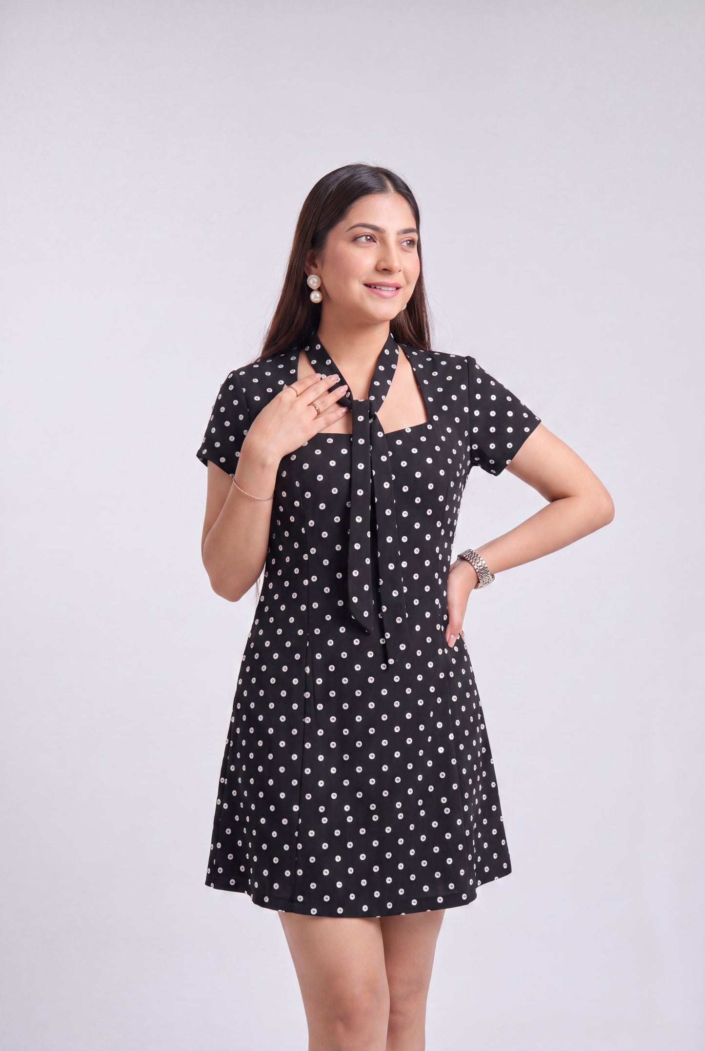 A woman modeling a short-sleeve black dress with a white polka dot pattern, featuring a tie-neck detail and a flared skirt, set against a plain light gray background.