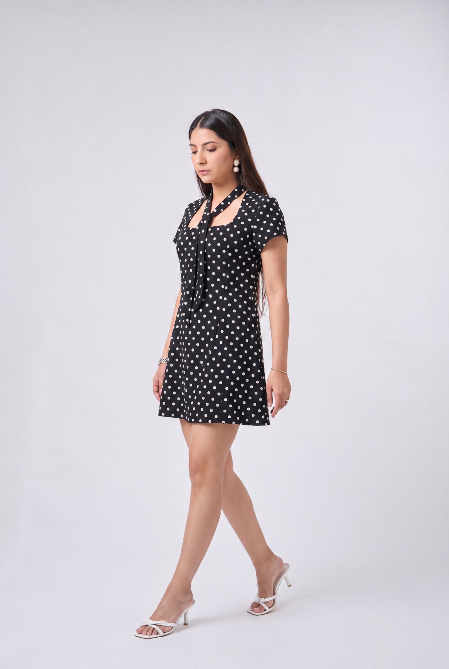 A woman modeling a short black dress with white polka dots, featuring a tie-neck detail and short sleeves, paired with white heeled sandals against a light gray studio background.