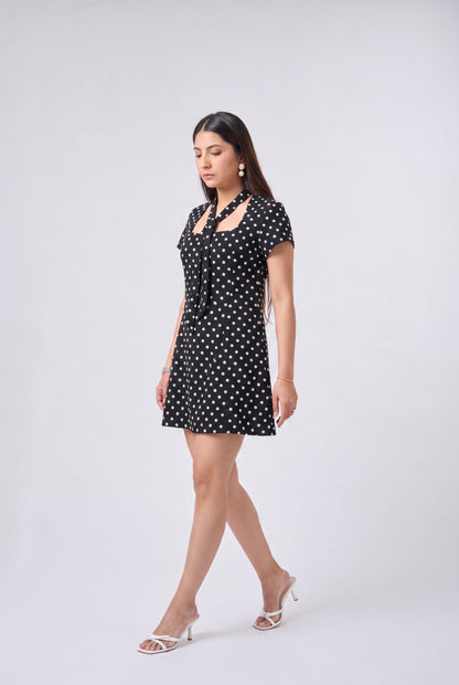 A woman modeling a short black dress with white polka dots, featuring a tie-neck detail and short sleeves, paired with white heeled sandals against a light gray studio background.