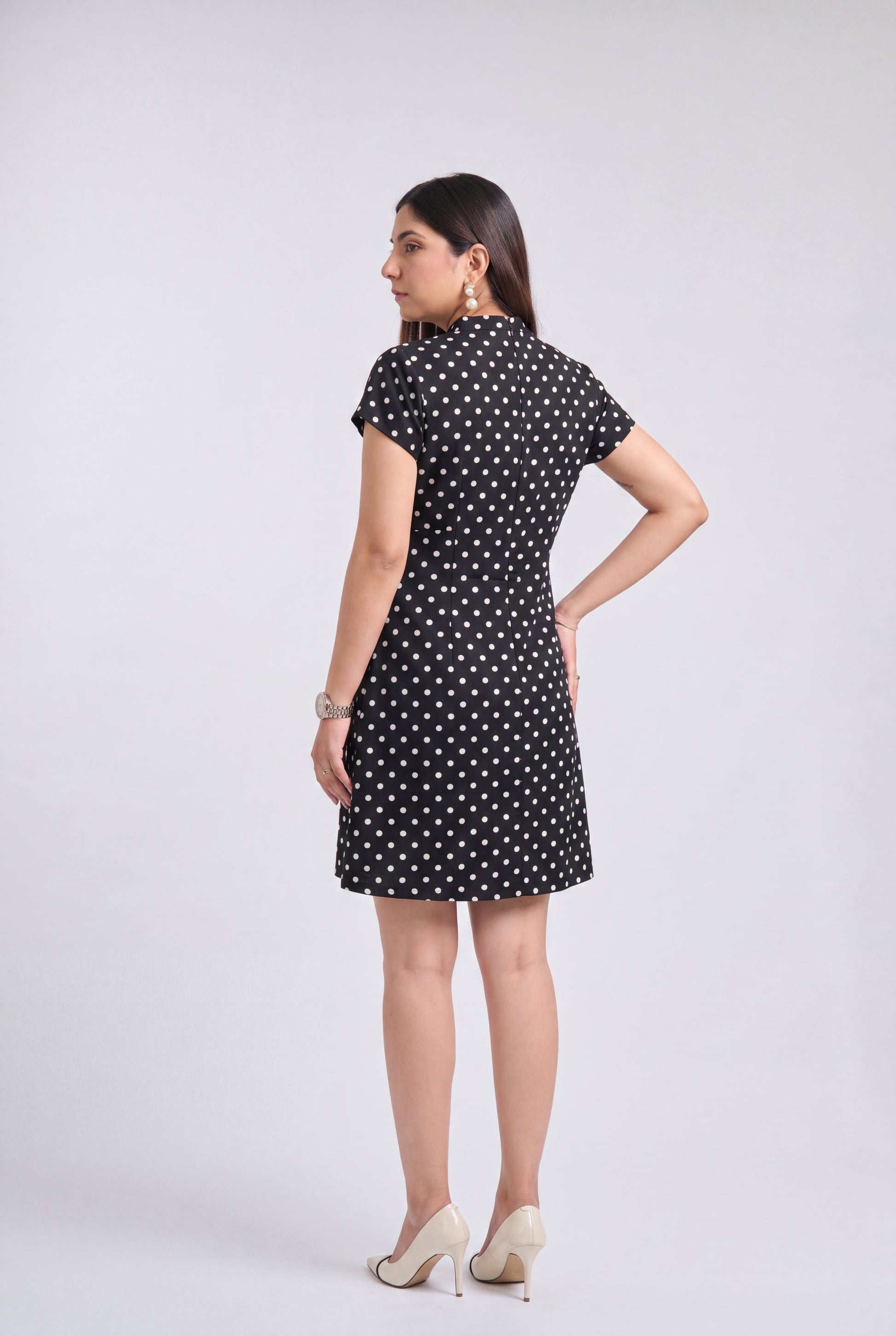 A woman modeling a short-sleeve black dress with a white polka dot pattern, shown from a rear three-quarter view against a light gray studio background.