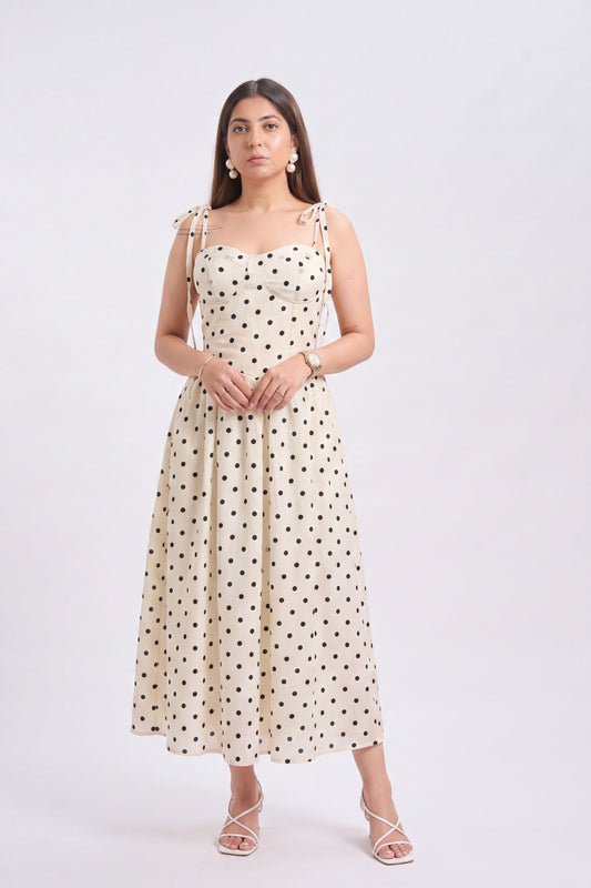 A woman modeling a sleeveless cream midi dress with black polka dots and tie-shoulder straps, paired with white strappy heels, against a plain white studio background.