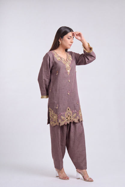 A woman modeling a traditional three-piece salwar kameez suit in a muted mauve-brown color with gold embroidery, standing against a light gray studio background.