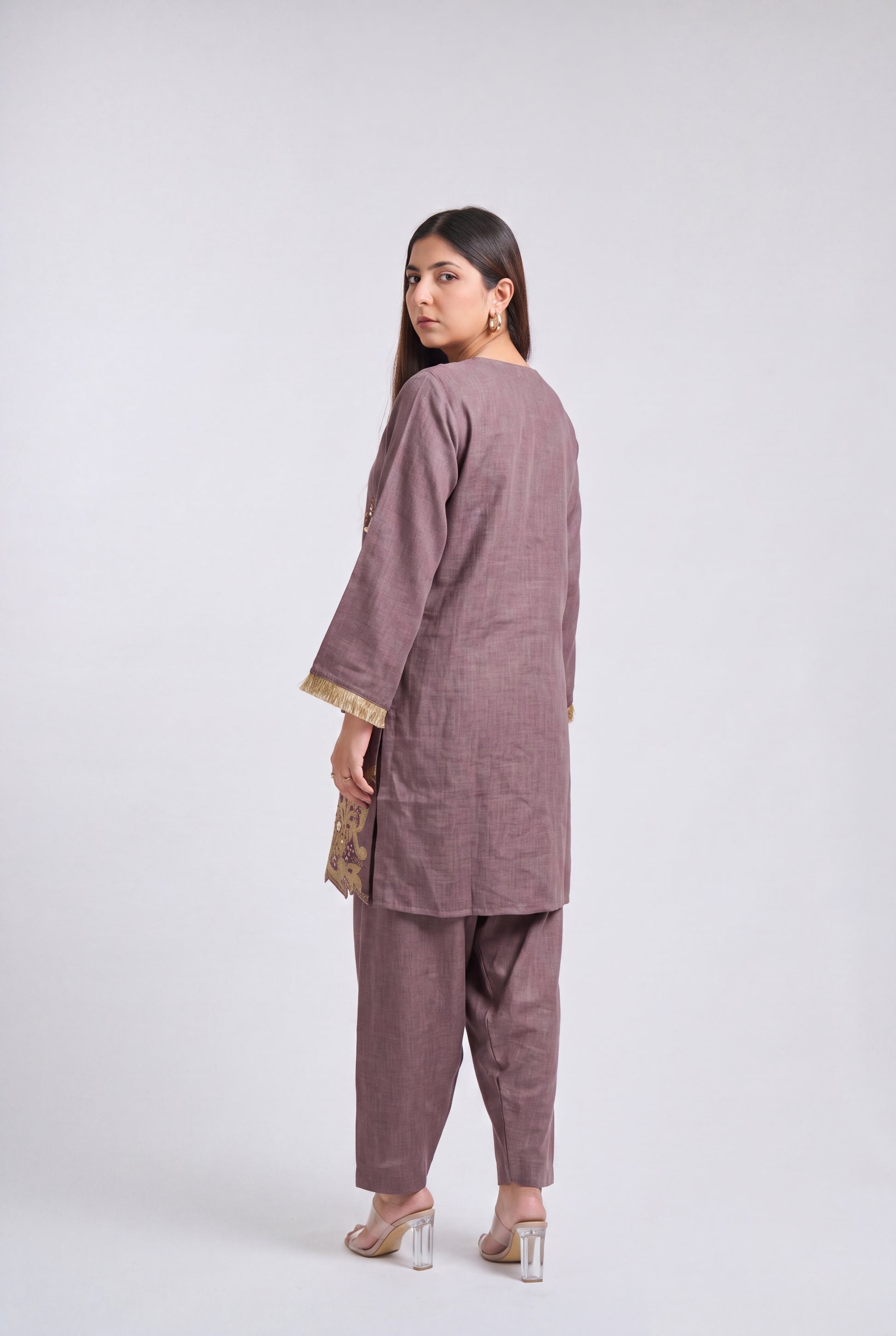 A woman modeling a two-piece dusty mauve kurta and trouser set, shown from a rear three-quarter angle against a light gray studio background.