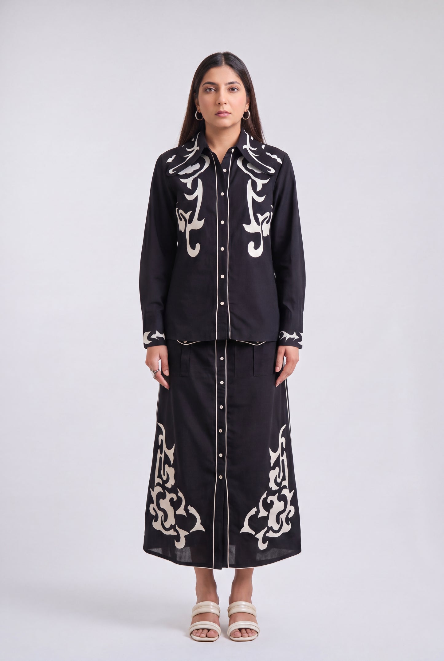A woman stands against a light gray studio background wearing a black long-sleeve button-front shirt and matching long skirt set with ornate white embroidered detailing.