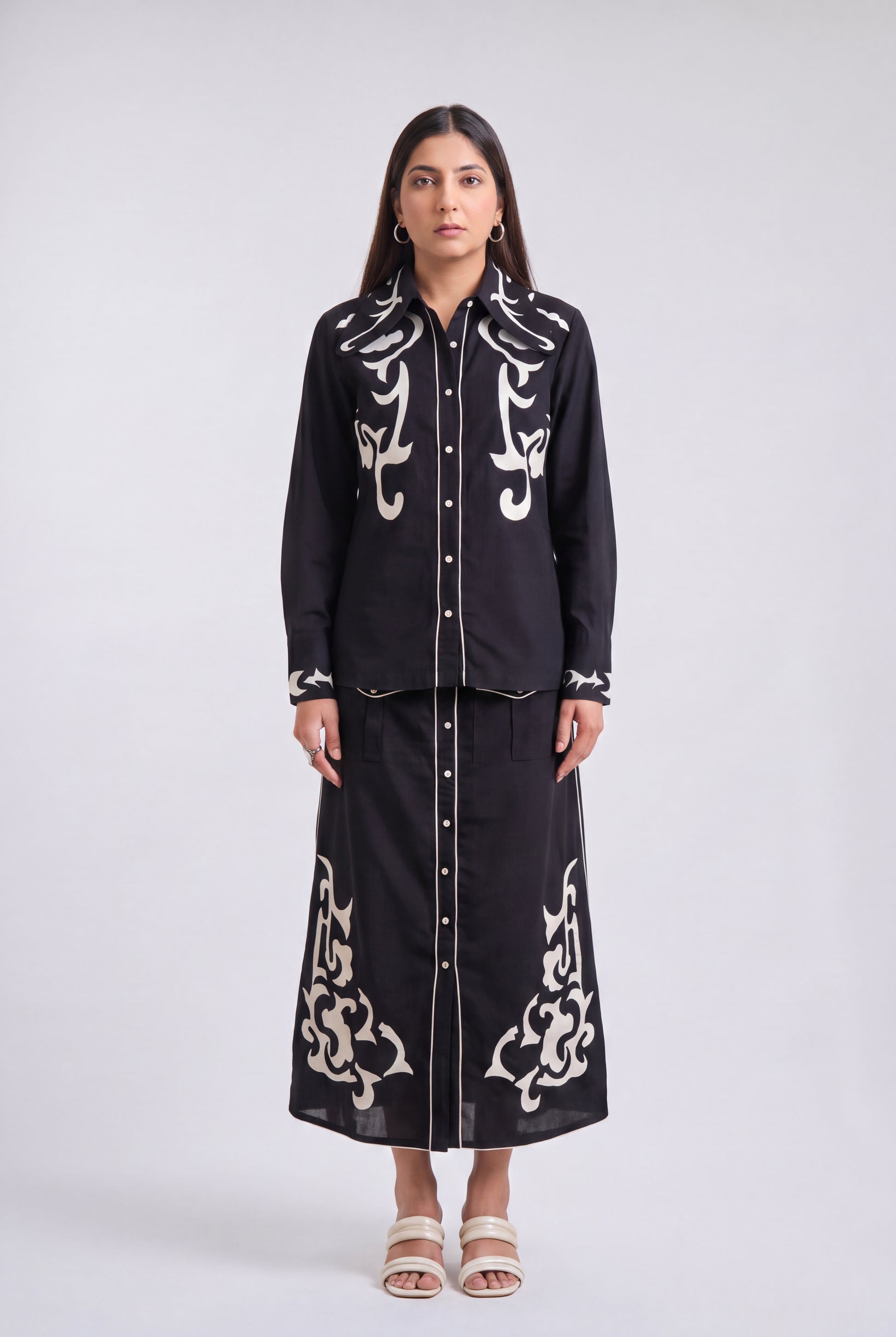 A woman stands against a light gray studio background wearing a black long-sleeve button-front shirt and matching long skirt set with ornate white embroidered detailing.