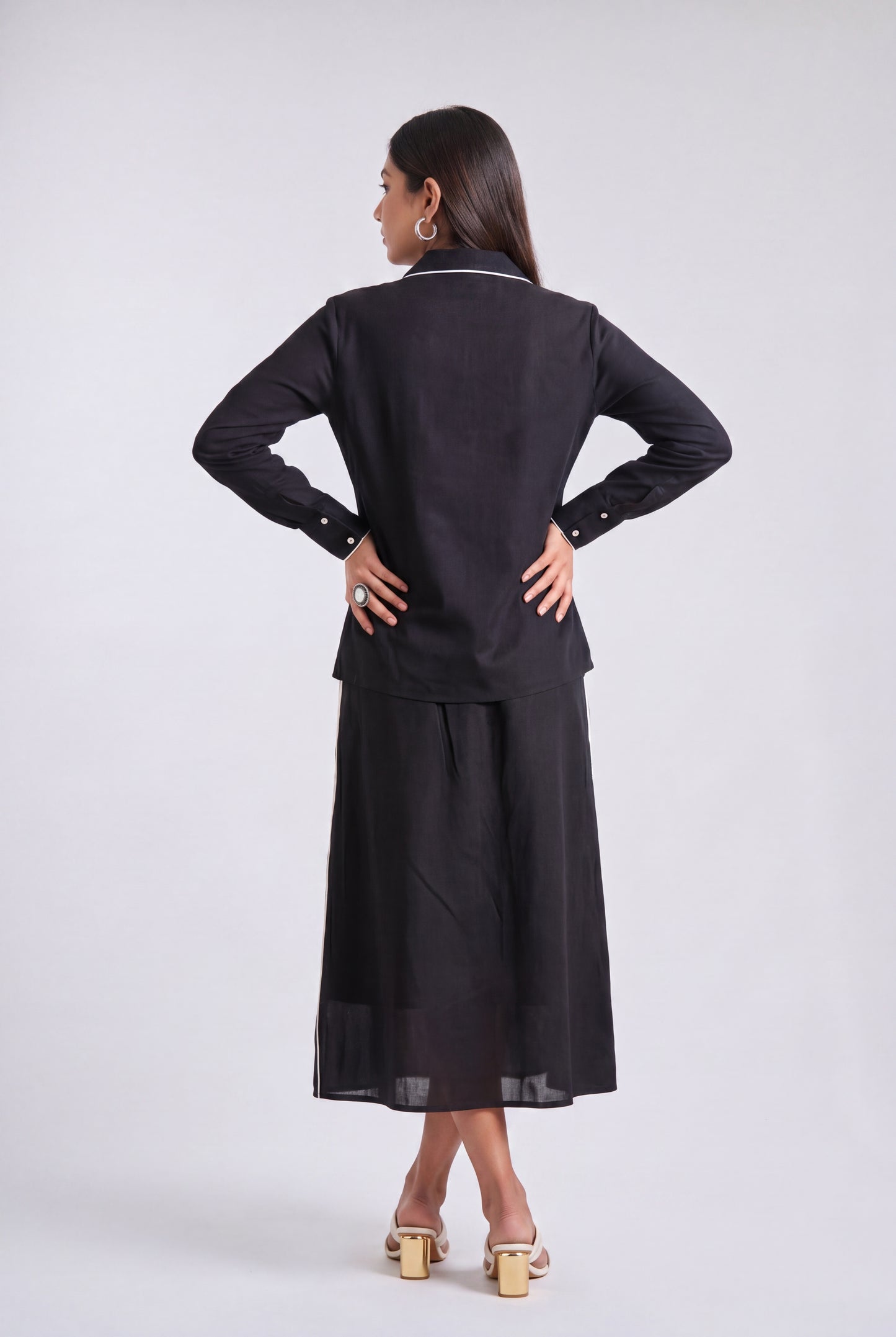 A woman is shown from the back wearing a black long-sleeve top and matching wide-leg skirt set, photographed against a plain light gray studio background.
