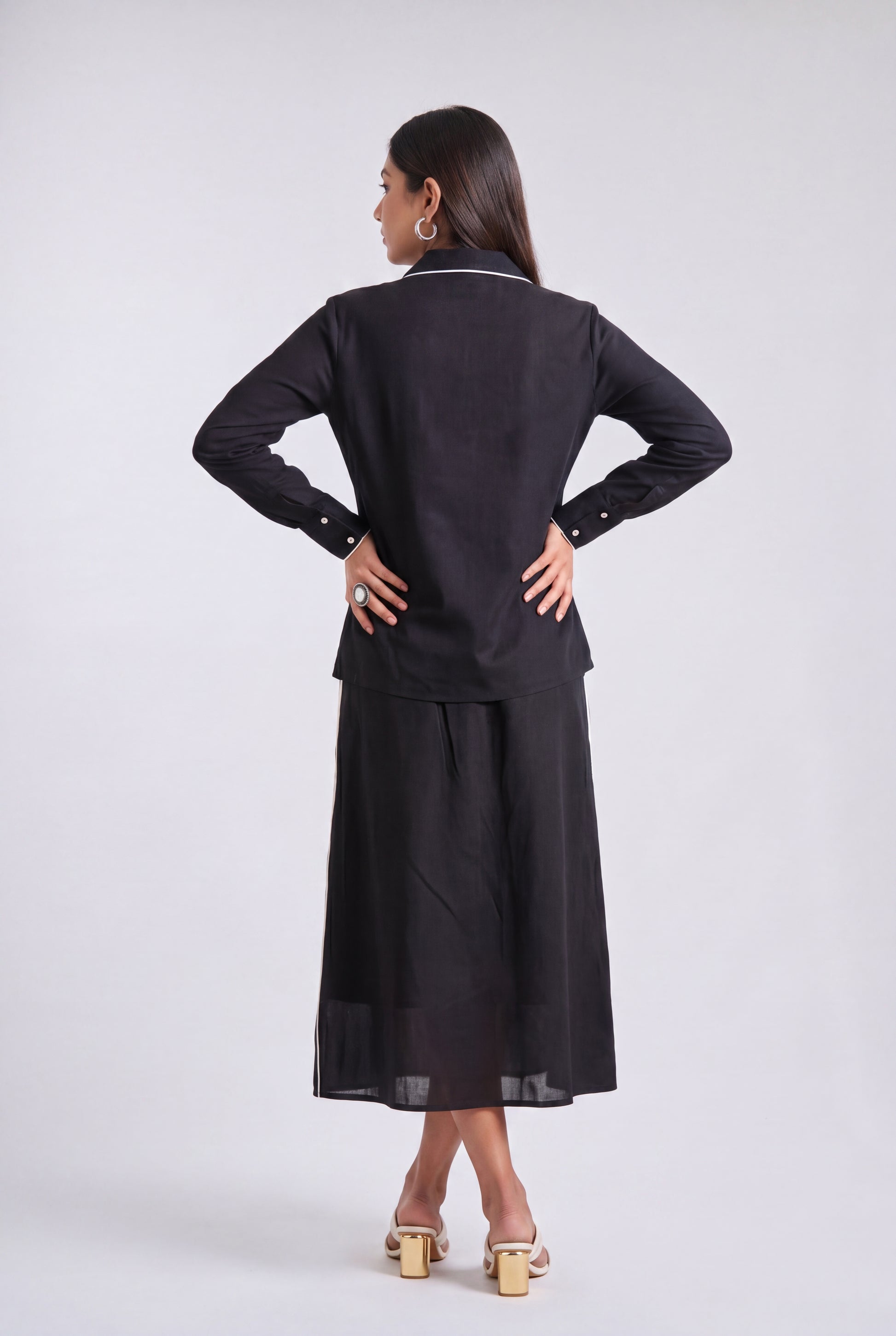 A woman is shown from the back wearing a black long-sleeve top and matching wide-leg skirt set, photographed against a plain light gray studio background.