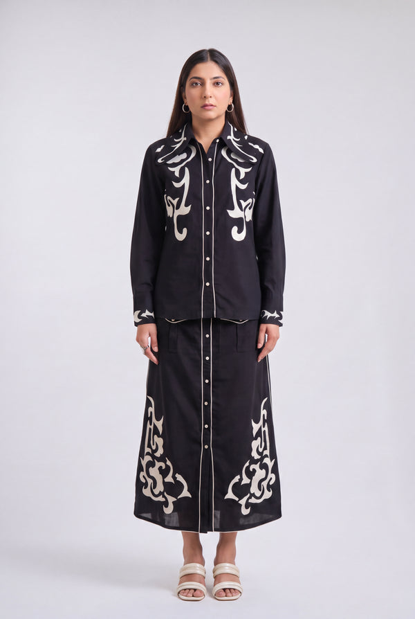 A woman stands against a light gray studio background wearing a black long-sleeve button-front shirt and matching long skirt set with ornate white embroidered detailing.