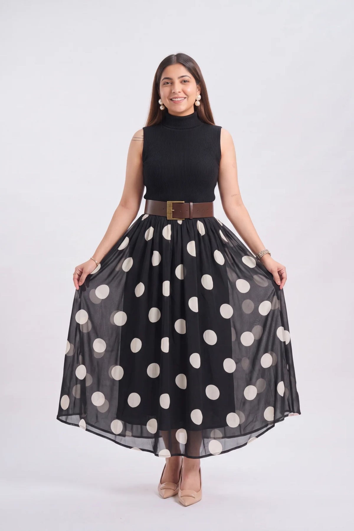 A woman modeling a sleeveless black mock-neck dress featuring a black and white polka dot skirt and a brown belt with a gold buckle, set against a plain light gray background.