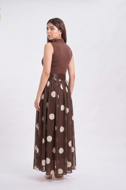 A woman modeling a sleeveless brown maxi dress with a high neck, a matching brown belt, and a brown skirt featuring large beige polka dots, shown from a rear three-quarter view against a plain white studio background.