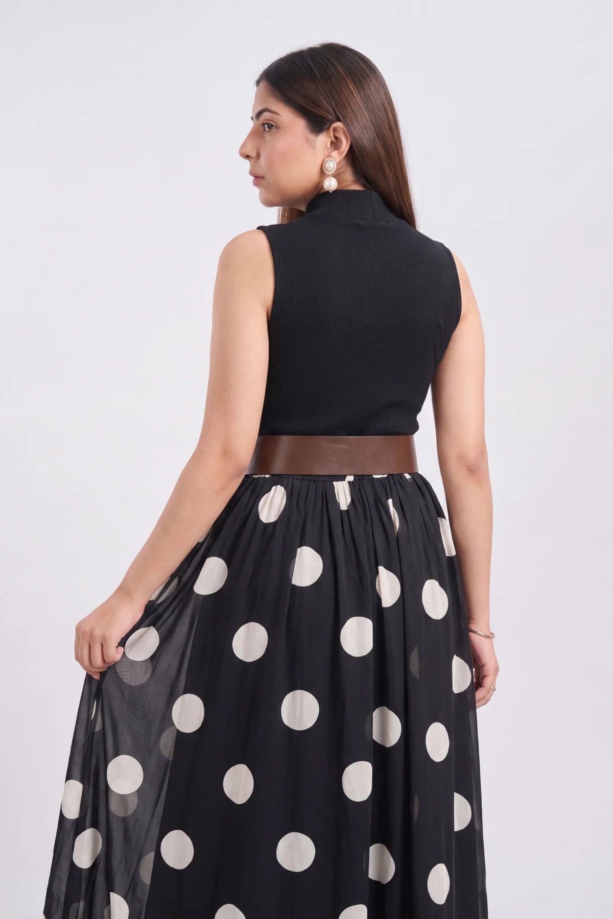 A woman shown from the back wearing a sleeveless black mock-neck top and a black midi skirt with large white polka dots, against a plain light gray studio background.
