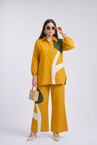 A woman modeling a matching two-piece outfit consisting of a mustard yellow button-up shirt and wide-leg trousers with abstract green and white graphic panels. She is wearing round sunglasses, holding a small woven handbag, and standing against a light gray studio background.