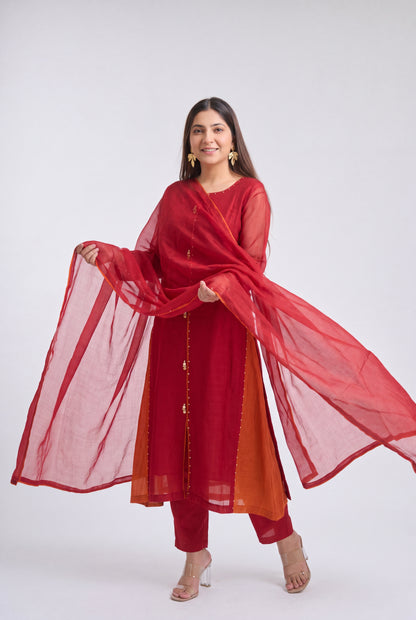 A woman modeling a red three-piece traditional outfit with a matching sheer dupatta, featuring a red kurta with gold button accents, red pants, and a rust-orange inner lining, set against a plain light gray studio background.