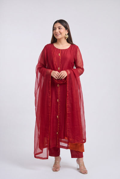A woman modeling a traditional Indian outfit consisting of a deep red kurta with gold button details, matching red pants, and a sheer red dupatta, set against a plain light gray background.
