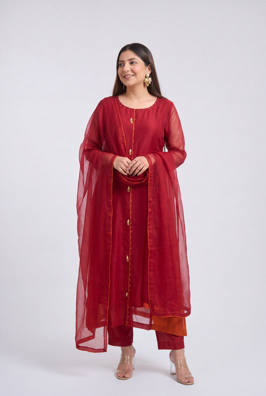 A woman modeling a traditional Indian outfit consisting of a deep red kurta with gold button details, matching red pants, and a sheer red dupatta, set against a plain light gray background.