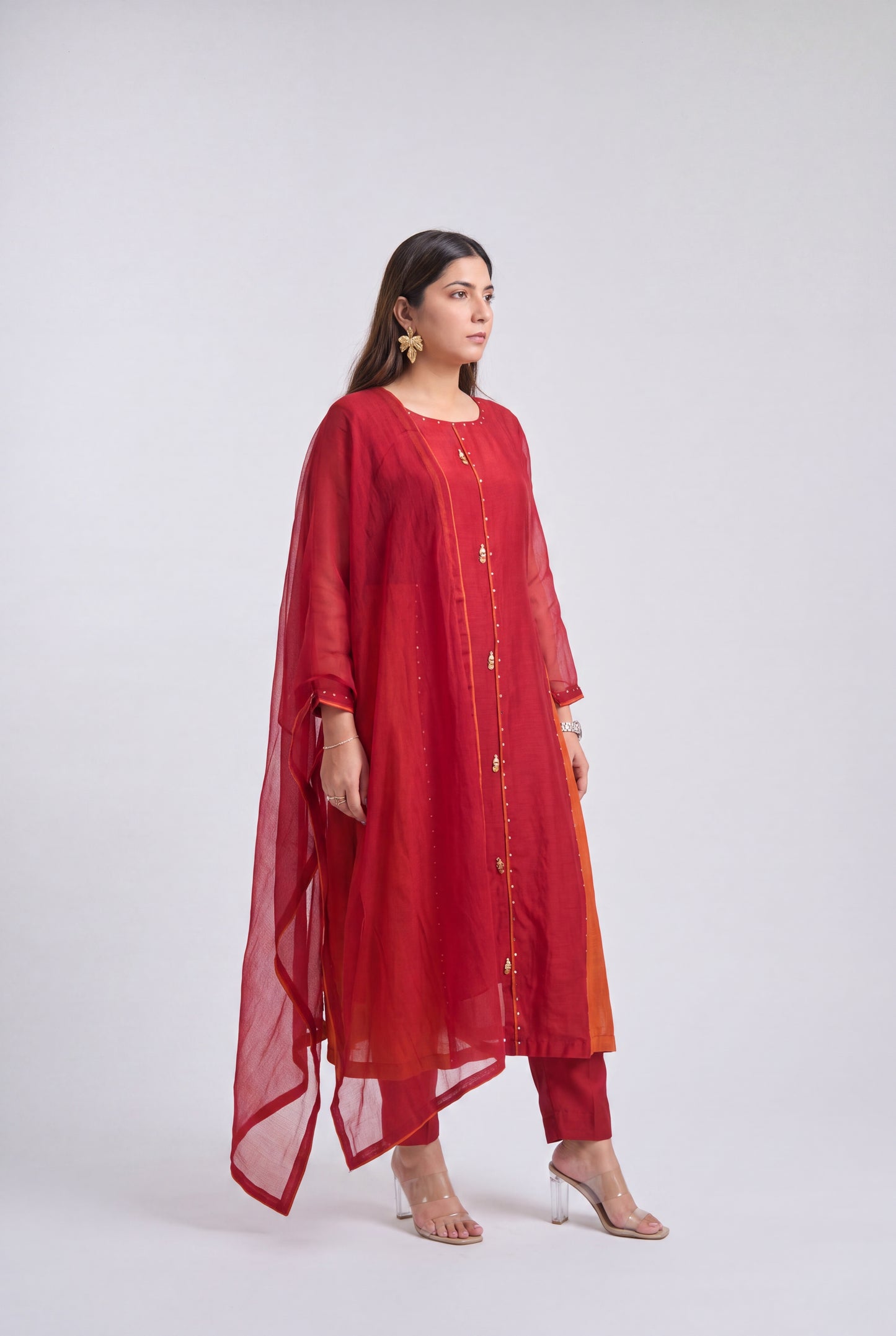 A woman modeling a red three-piece traditional outfit consisting of a long kurta with gold button details, matching pants, and a sheer red dupatta, set against a plain light gray studio background.