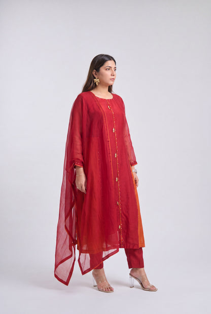 A woman modeling a red three-piece traditional outfit consisting of a long kurta with gold button details, matching pants, and a sheer red dupatta, set against a plain light gray studio background.