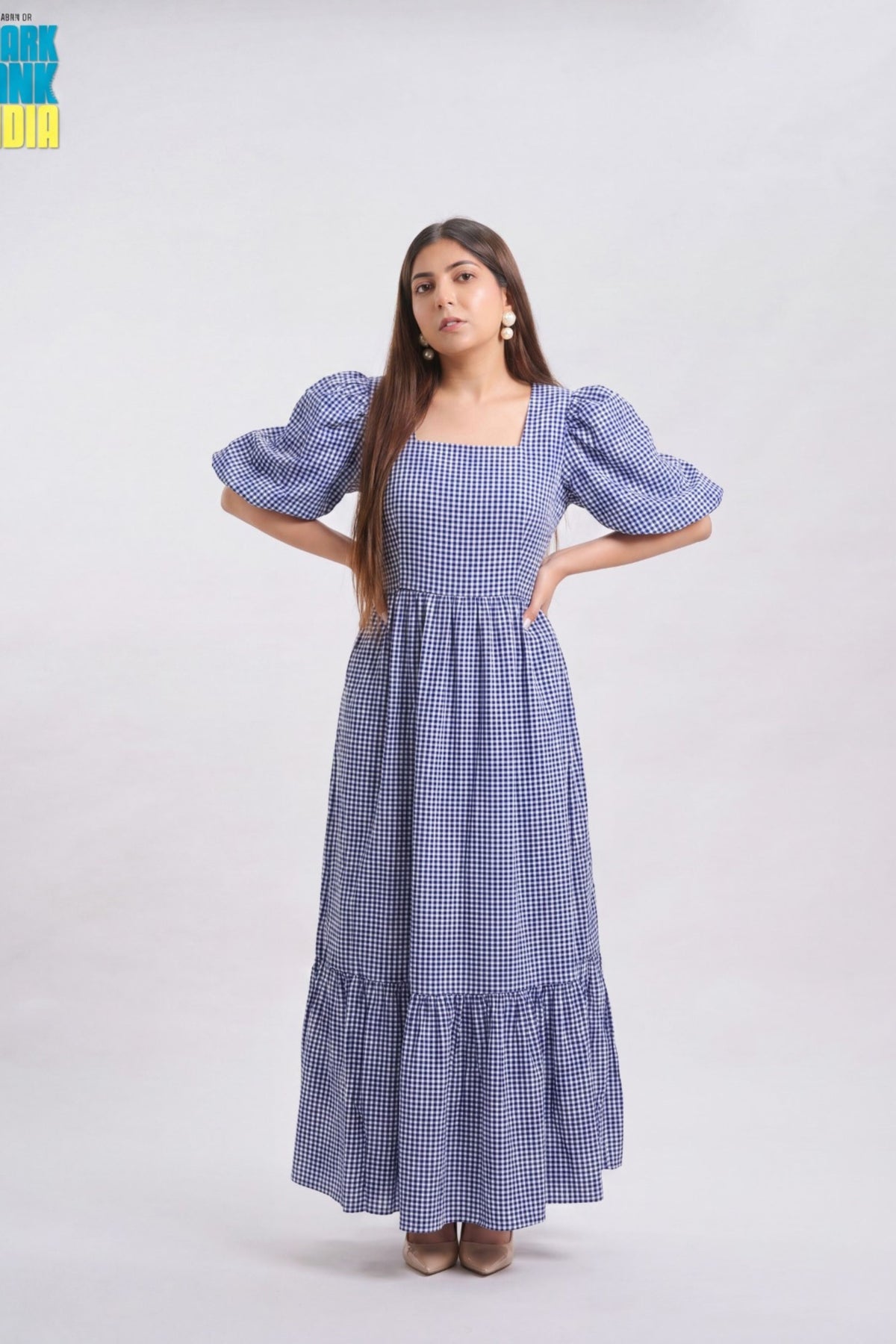 A woman modeling a blue and white gingham maxi dress with puff sleeves, a square neckline, and a tiered skirt, standing against a light gray studio background.