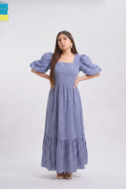 A woman modeling a blue and white gingham maxi dress with puff sleeves, a square neckline, and a tiered skirt, standing against a light gray studio background.