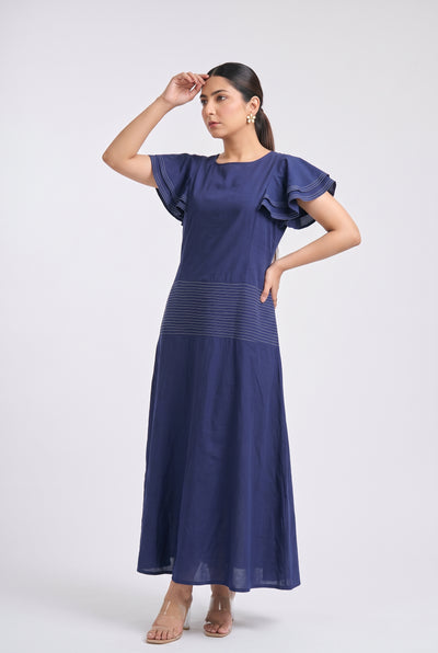 A woman modeling a navy blue maxi dress with ruffled short sleeves and a subtle horizontal textured stripe detail at the waist, posed against a light gray studio background.