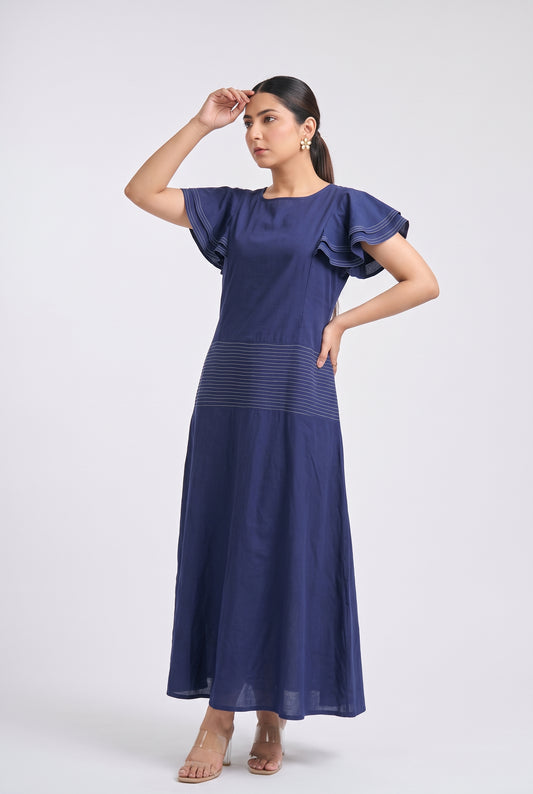 A woman modeling a navy blue maxi dress with ruffled short sleeves and a subtle horizontal textured stripe detail at the waist, posed against a light gray studio background.