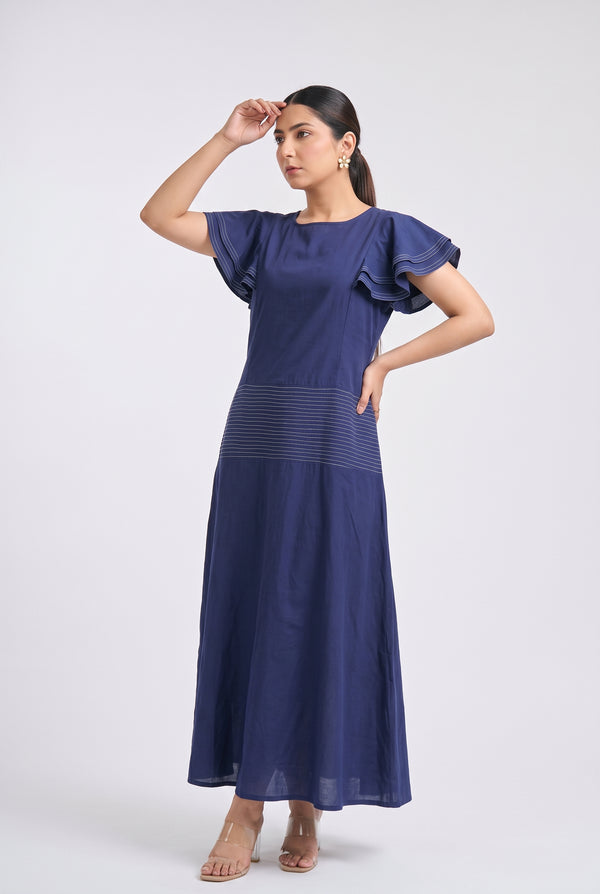A woman modeling a navy blue maxi dress with ruffled short sleeves and a subtle horizontal textured stripe detail at the waist, posed against a light gray studio background.