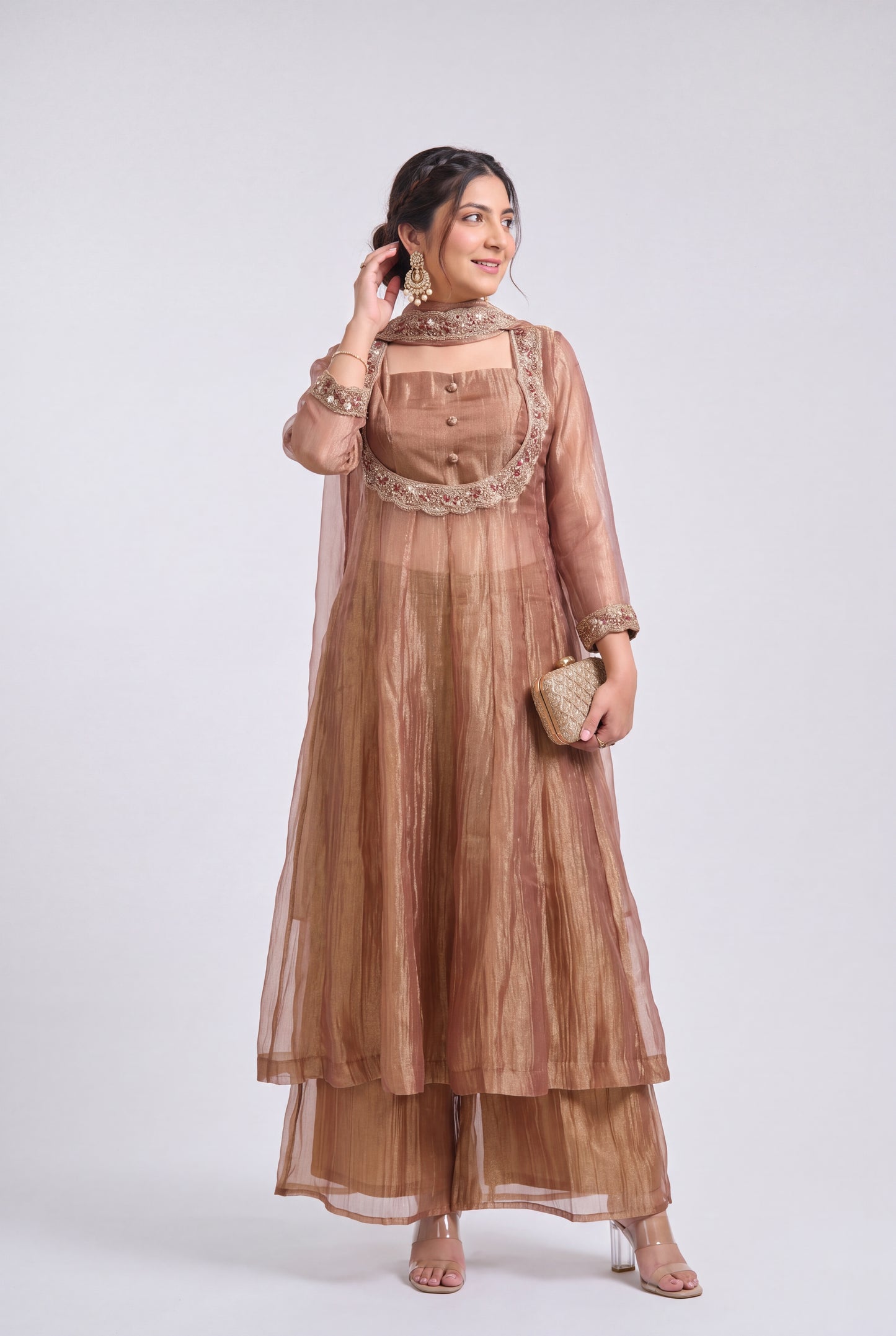 A woman models a traditional three-piece salwar kameez set in a dusty rose-brown color with intricate gold embroidery. The set includes a long kurta with a high-neck collar, matching wide-leg pants, and a sheer dupatta.