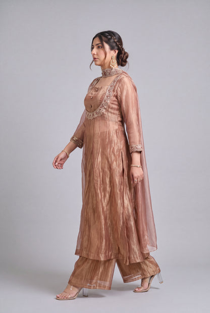 A woman modeling a traditional three-piece salwar kameez suit in a dusty rose-brown color with gold embroidery, paired with matching wide-leg trousers and a sheer matching dupatta, set against a solid gray studio background.