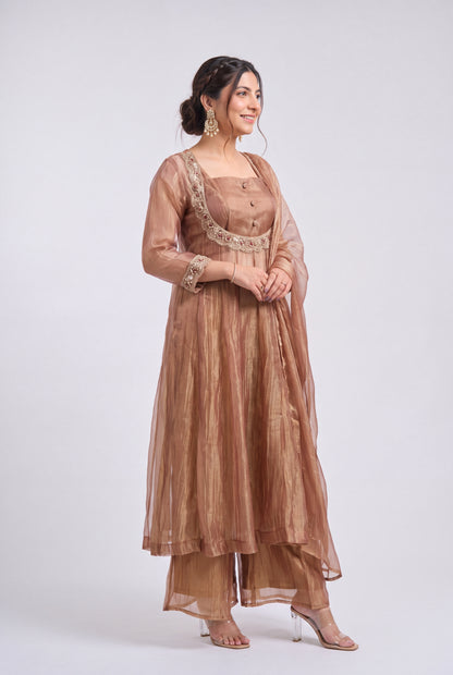 A woman viewed from the back wearing a metallic bronze traditional outfit with a long kurta and matching pants, standing against a light gray studio background.