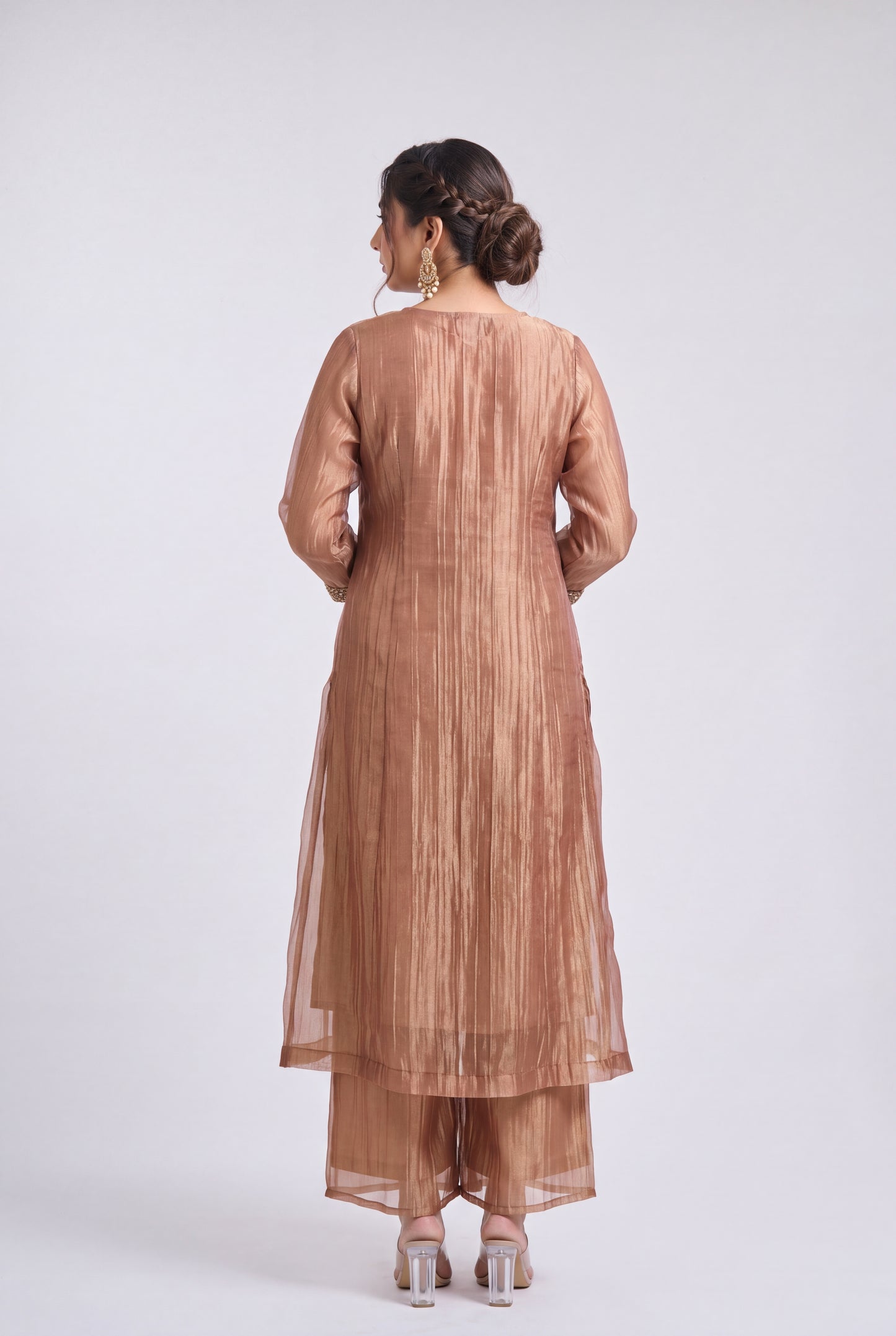 A woman viewed from the back wearing a metallic bronze traditional outfit with a long kurta and matching pants, standing against a light gray studio background.