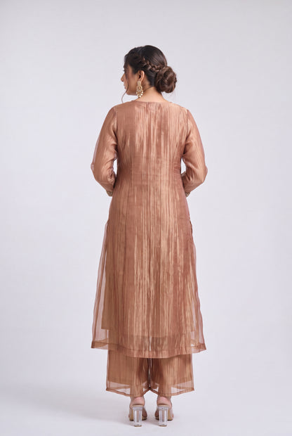A woman viewed from the back wearing a metallic bronze traditional outfit with a long kurta and matching pants, standing against a light gray studio background.