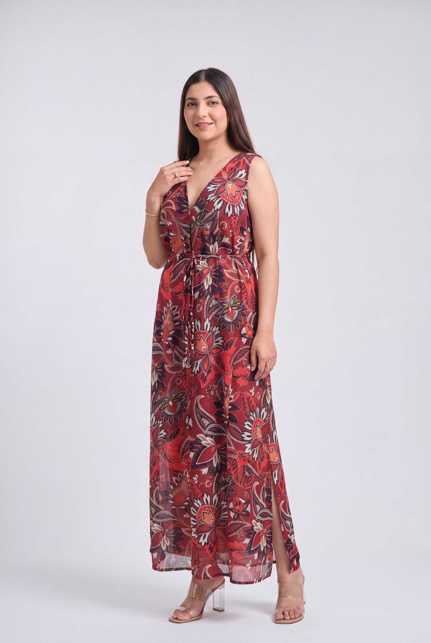 A woman modeling a sleeveless, floor-length maxi dress with a red, black, and beige floral pattern against a light gray studio background.