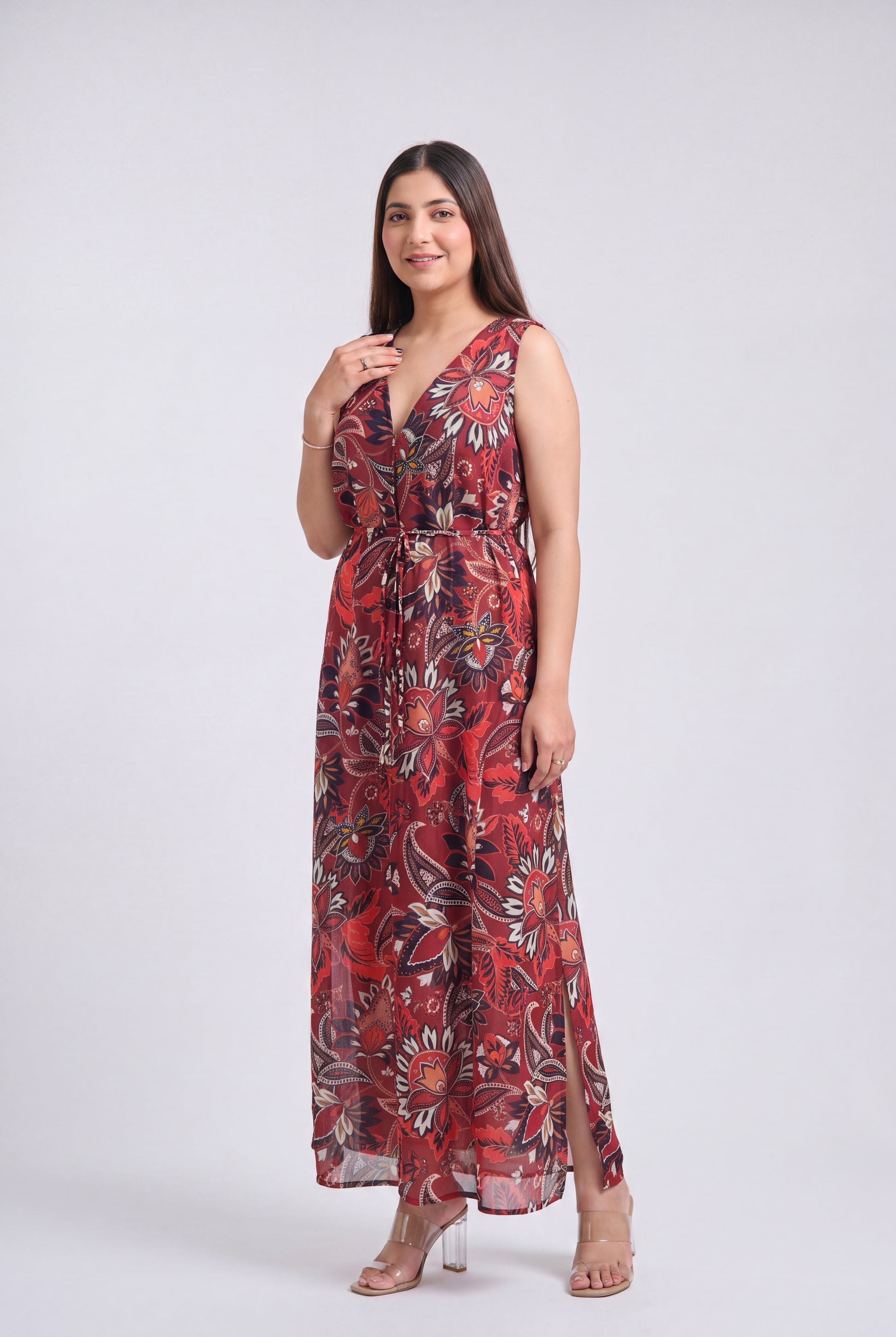 A woman modeling a sleeveless, floor-length maxi dress with a red, black, and beige floral pattern against a light gray studio background.