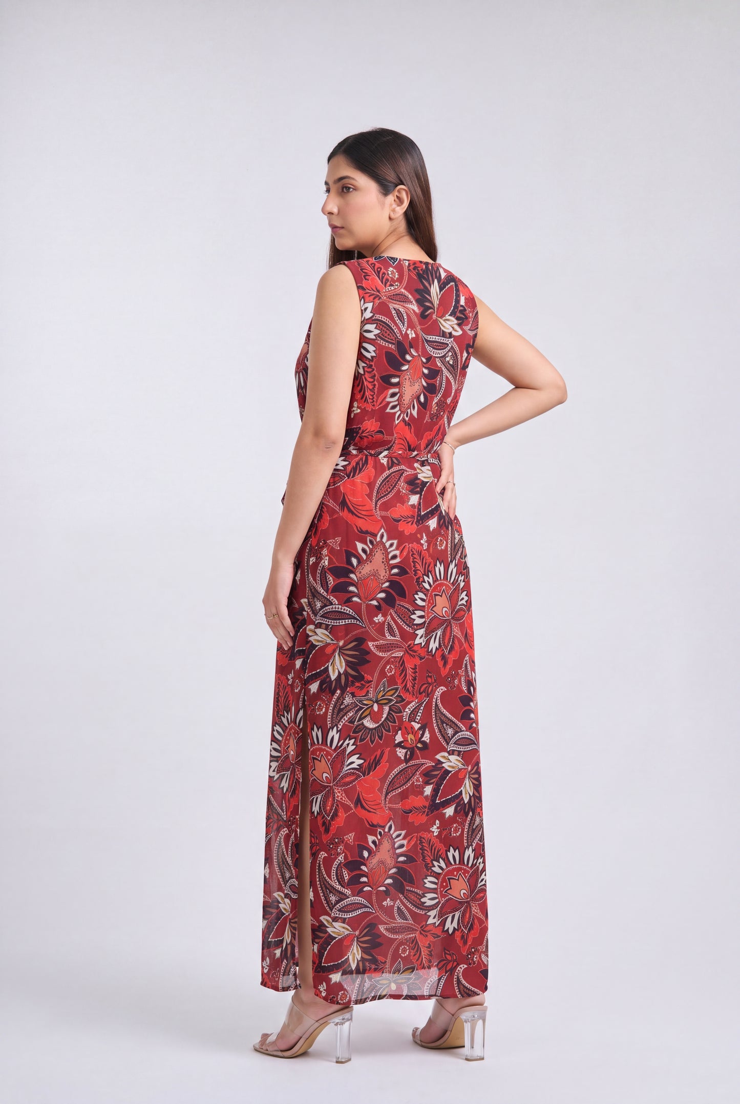 A woman modeling a sleeveless, floor-length maxi dress with a red, black, and beige floral pattern against a light gray studio background.