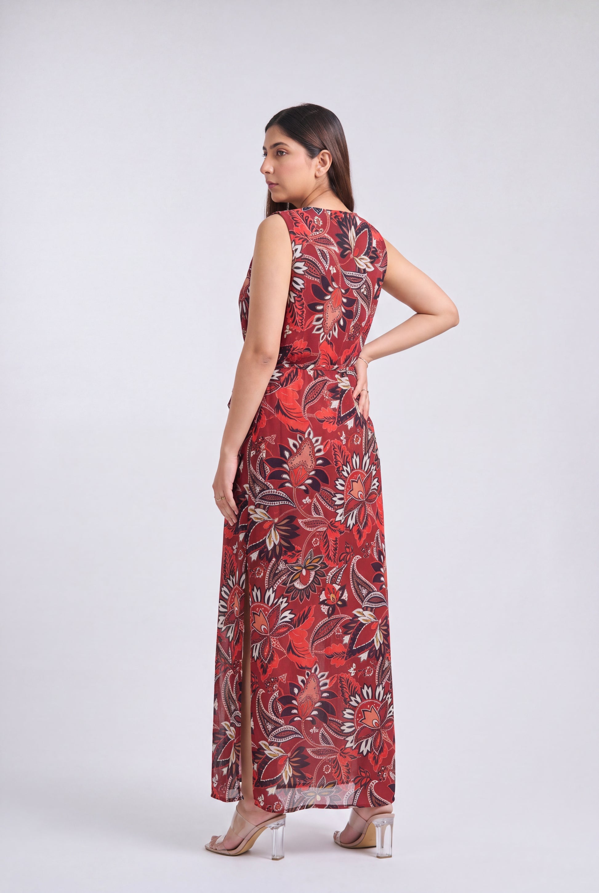 A woman modeling a sleeveless, floor-length maxi dress with a red, black, and beige floral pattern against a light gray studio background.