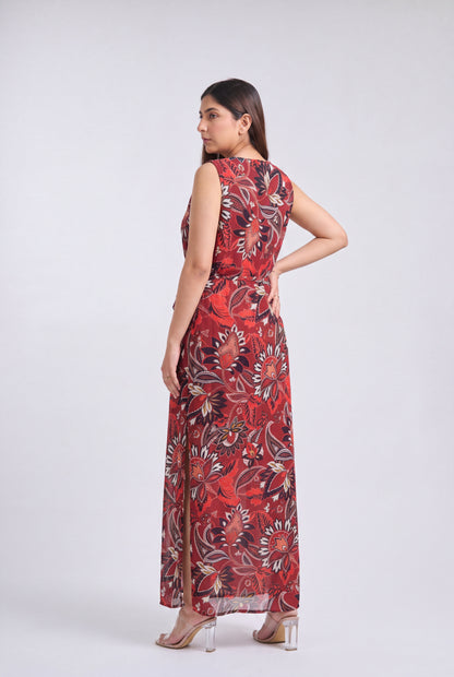 A woman modeling a sleeveless, floor-length maxi dress with a red, black, and beige floral pattern against a light gray studio background.