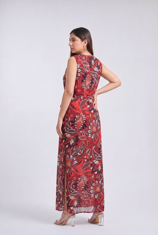 A woman modeling a sleeveless, floor-length maxi dress with a red, black, and beige floral pattern against a light gray studio background.