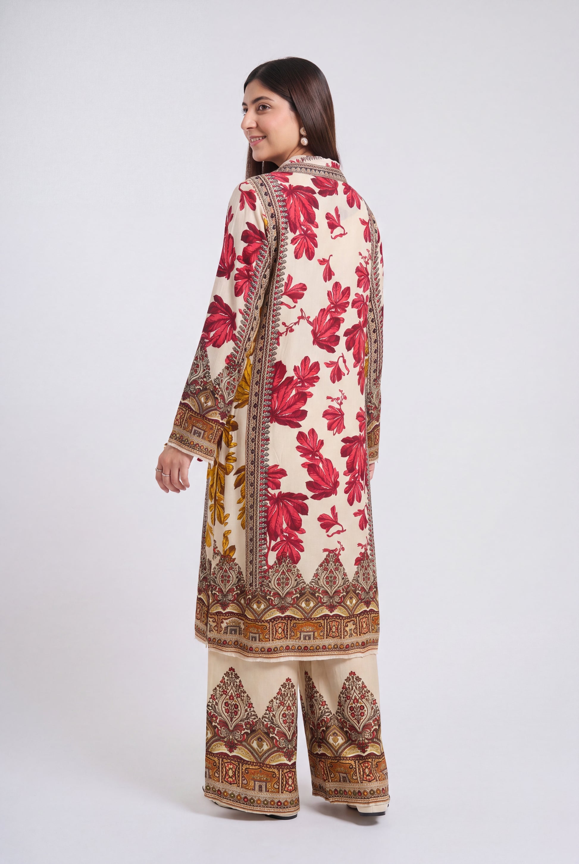 A woman modeling a traditional South Asian outfit, viewed from the back. The outfit consists of a long cream-colored kurta with a bold red floral print and ornate brown borders, paired with matching wide-leg trousers.