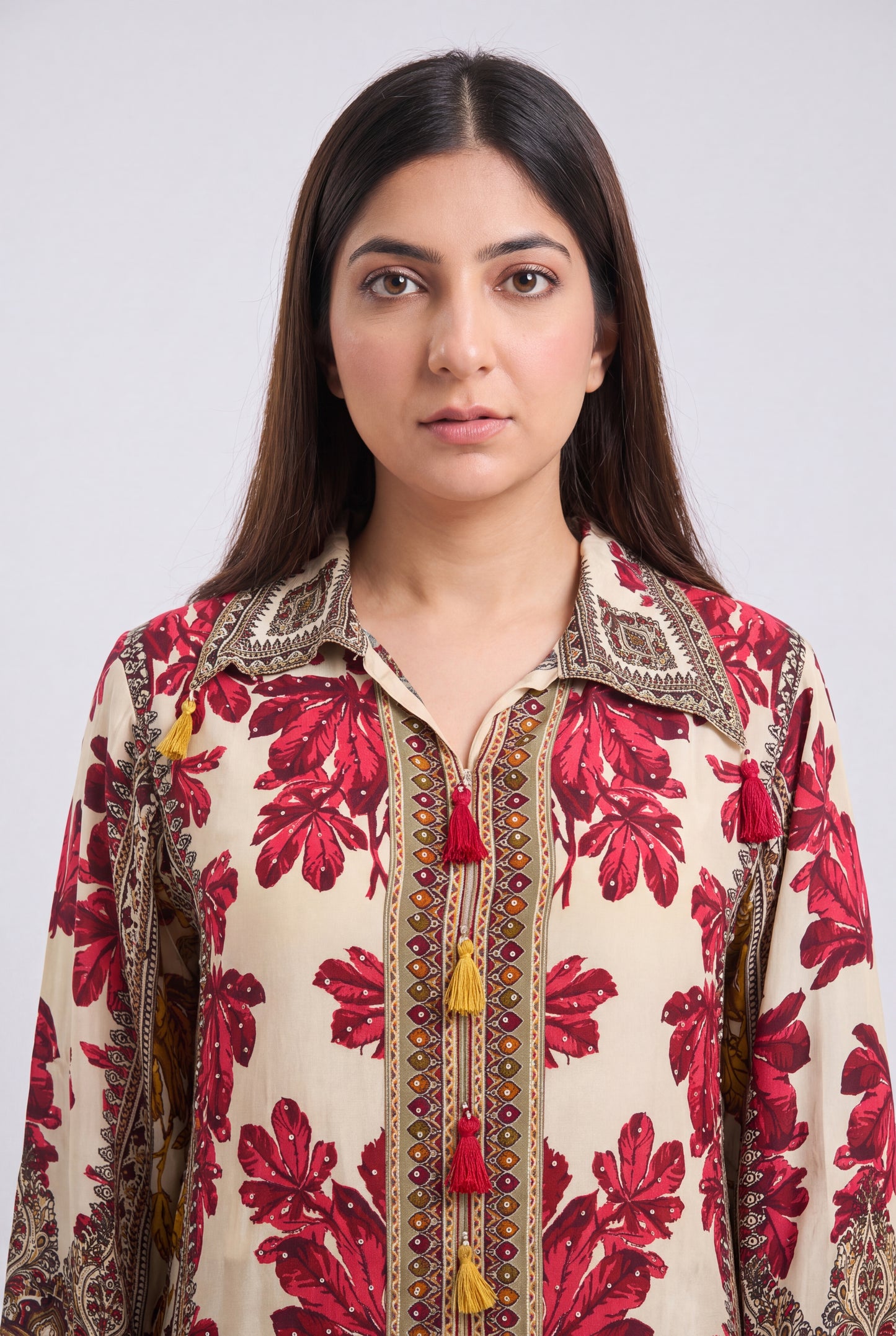 A woman wearing a cream-colored long-sleeve shirt with a red floral print and ornate brown embroidery with tassel details, set against a plain light gray background.