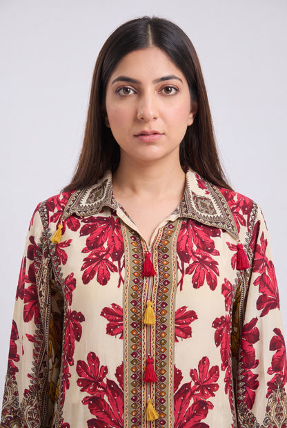 A woman wearing a cream-colored long-sleeve shirt with a red floral print and ornate brown embroidery with tassel details, set against a plain light gray background.