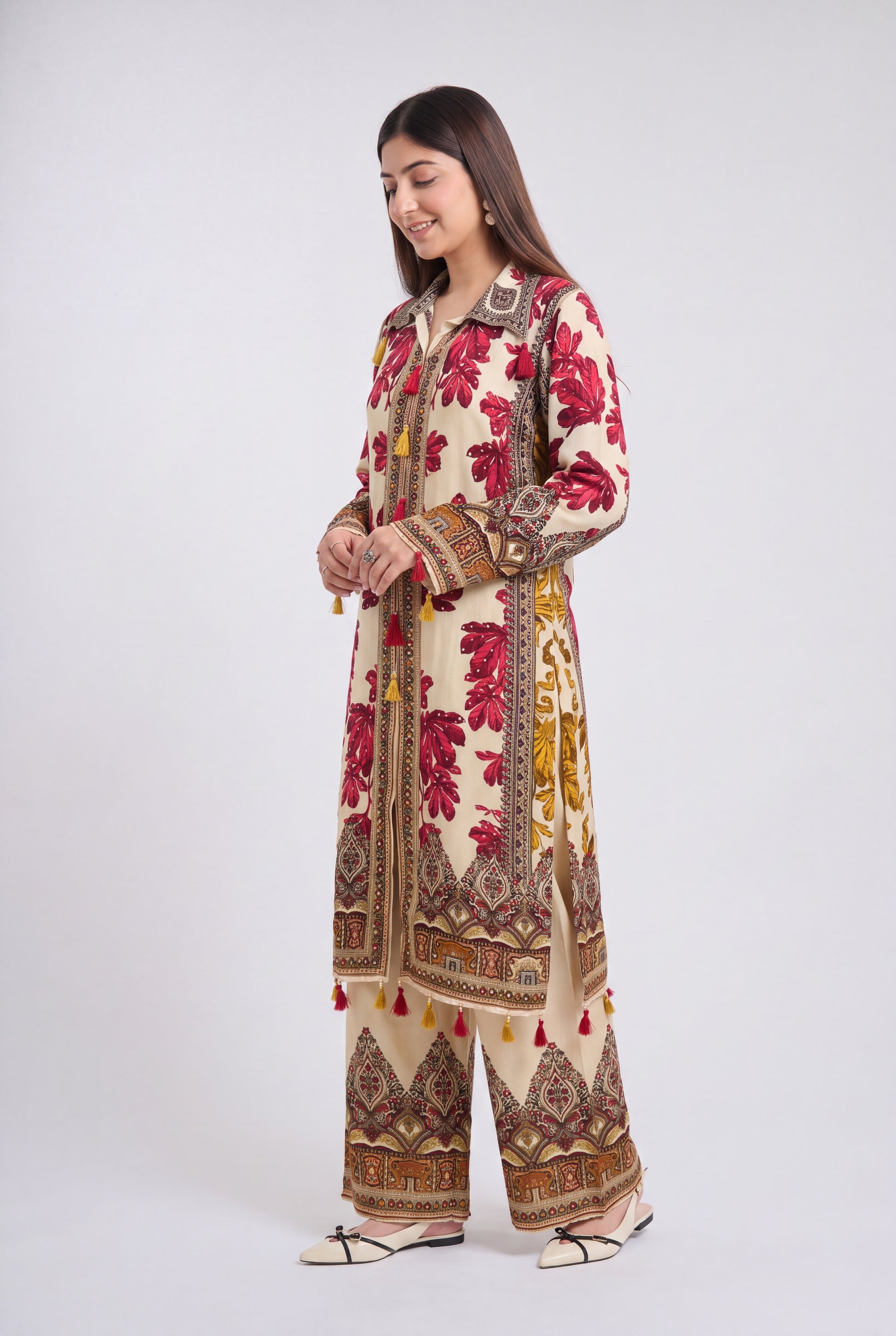 A woman modeling a traditional South Asian outfit consisting of a long cream-colored kurta and matching wide-leg trousers, decorated with red floral patterns and brown geometric borders, set against a plain light gray background.