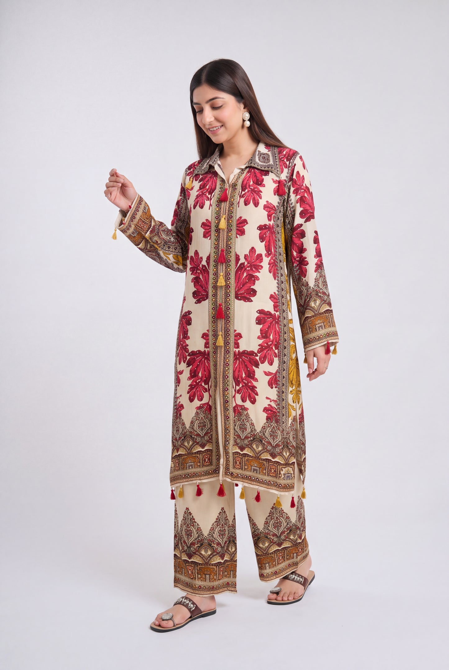A woman modeling a traditional South Asian outfit consisting of a long cream-colored kurta with red floral embroidery and brown patterns, paired with matching wide-leg trousers, against a light gray studio background.